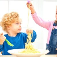 two kids eating plain spaghetti from a bowl