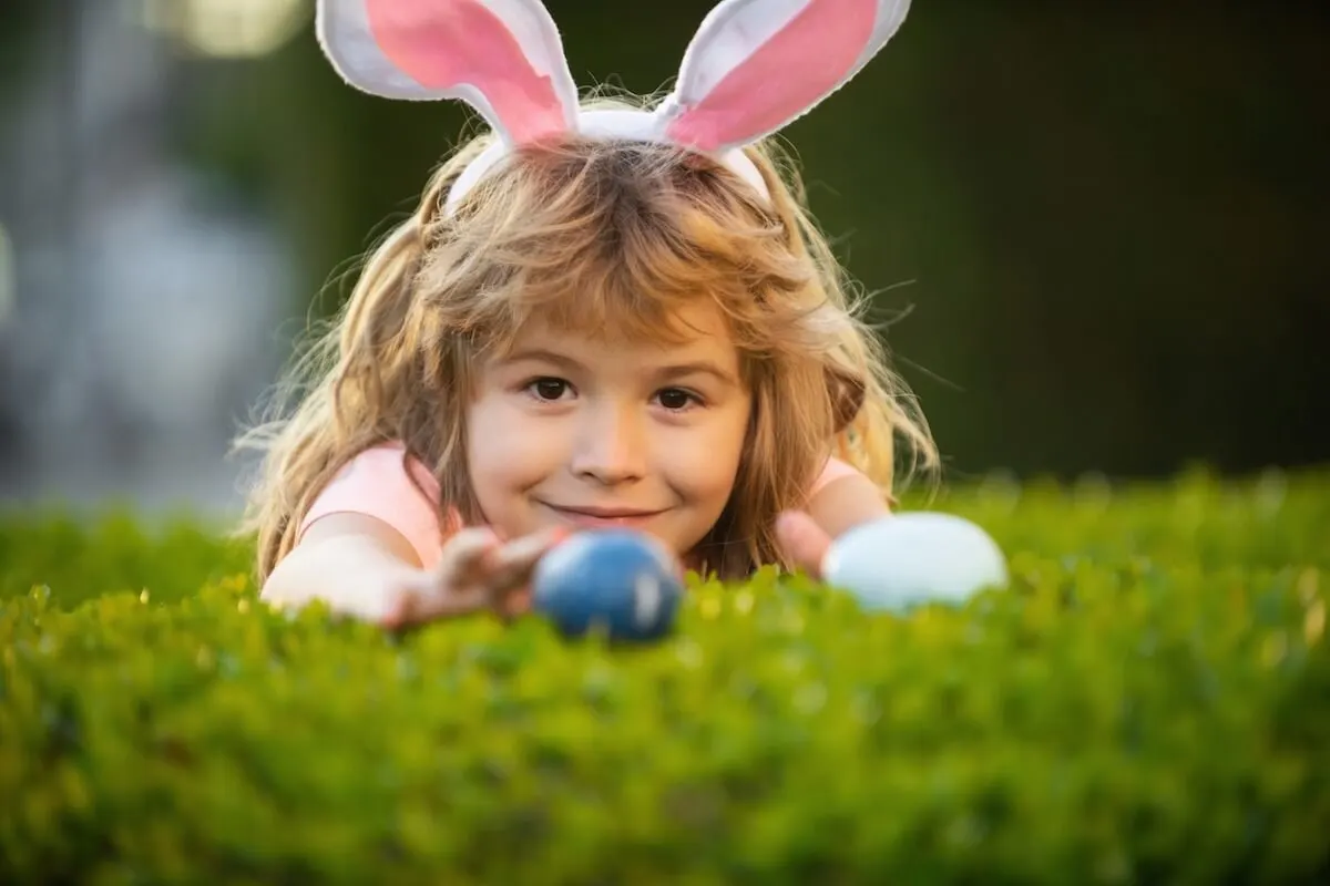 Child wearing bunny ears finding easter egg on top of a hedge