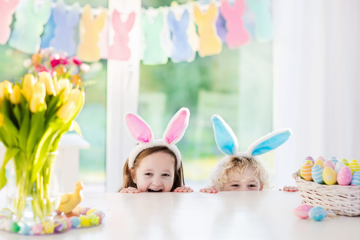 Two kids wearing bunny ears peering over a kitchen counter at easter eggs