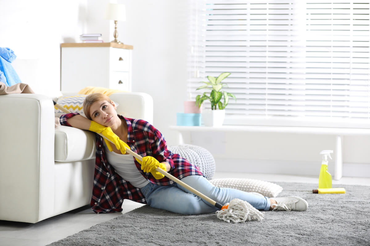 Woman feeling overwhelmed by cleaning her house, sitting on lounge room floor with mop