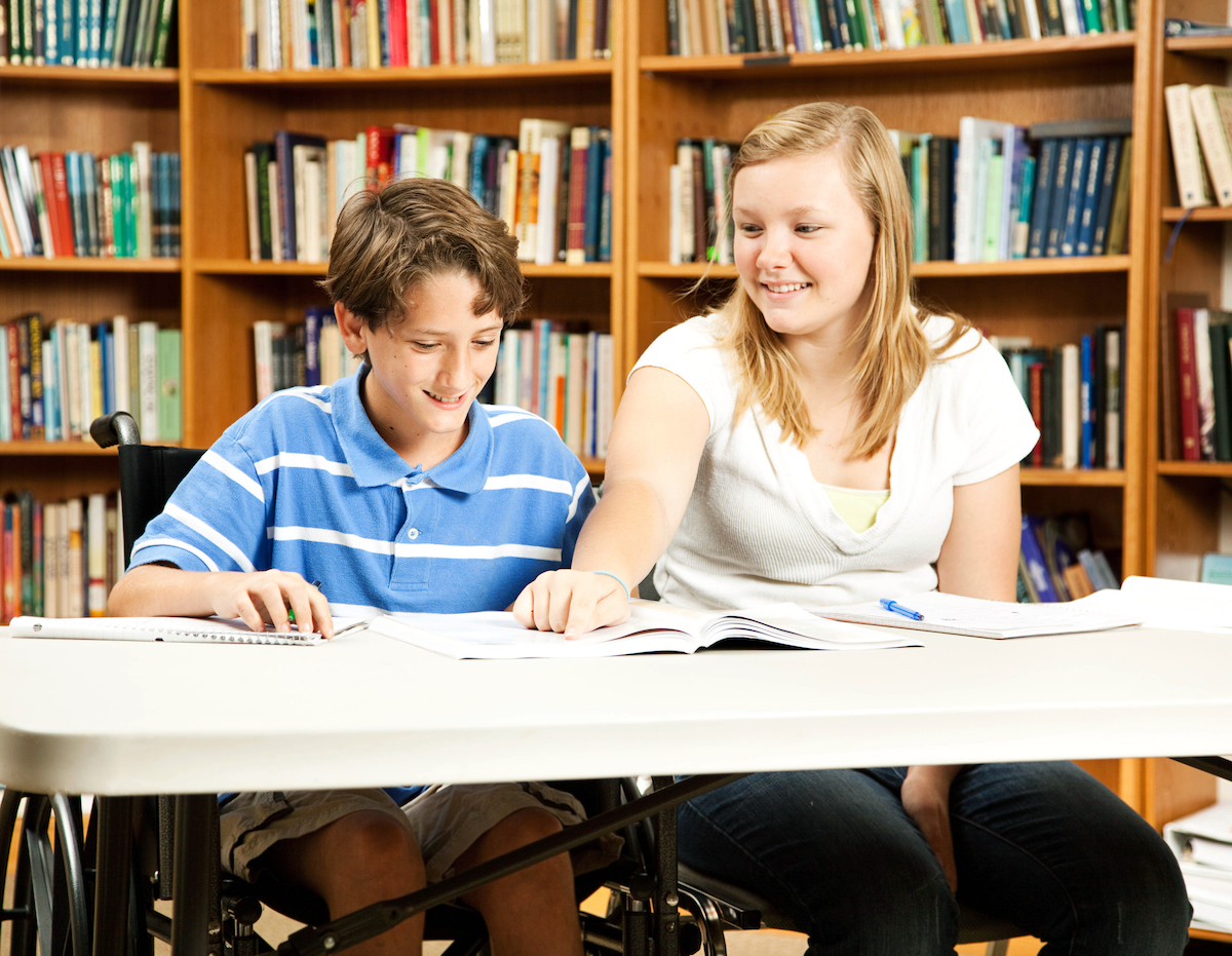 Teenage girl mentoring a younger, disabled boy in the school library.