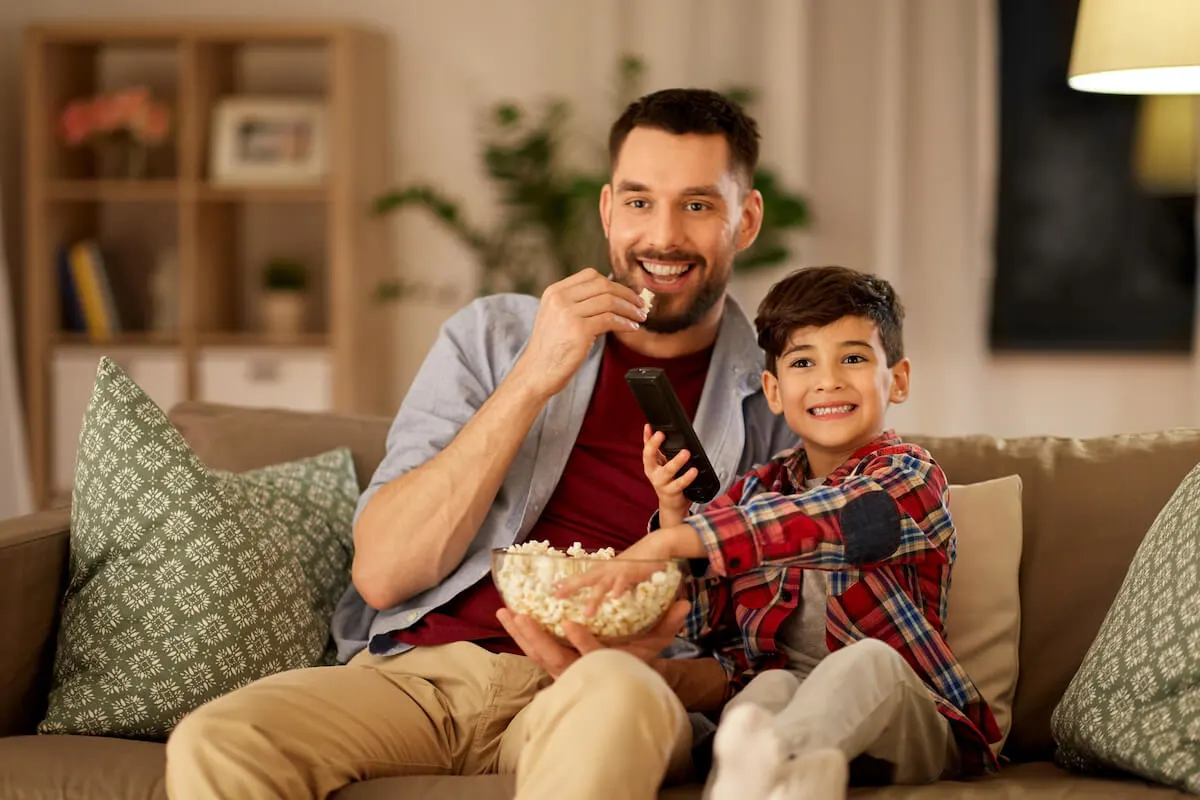 Young son sitting with father on couch with a bowl of popcorn and the tv remote