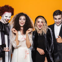 four people dressed up in halloween costumes including a clown, bride, witch and vampire with an orange background