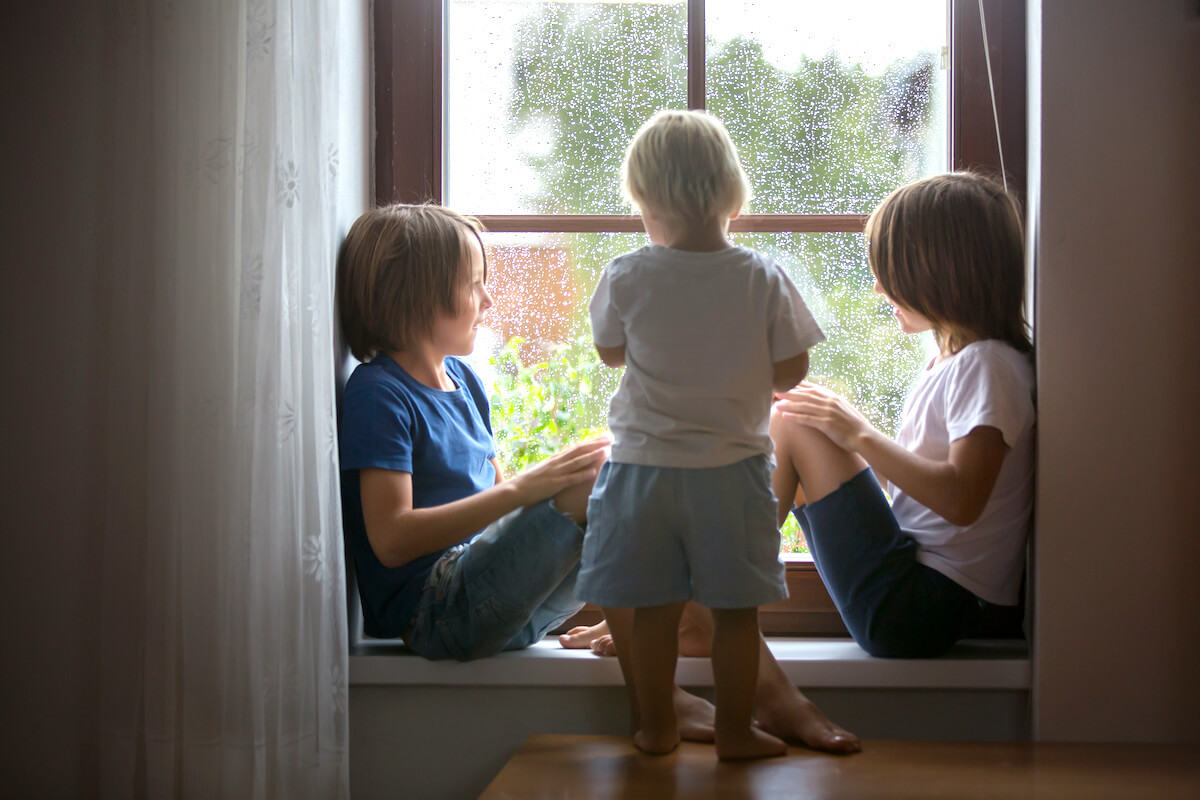 Three young boys sitting on the window seat inside their house looking out at the rain