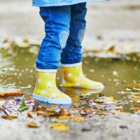 Small child wearing jeans and yellow gumboots jumping in puddles on a rainy day