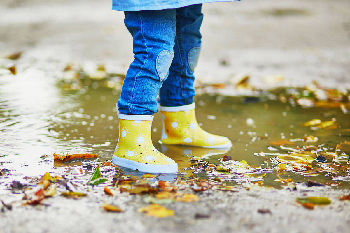 Small child wearing jeans and yellow gumboots jumping in puddles on a rainy day