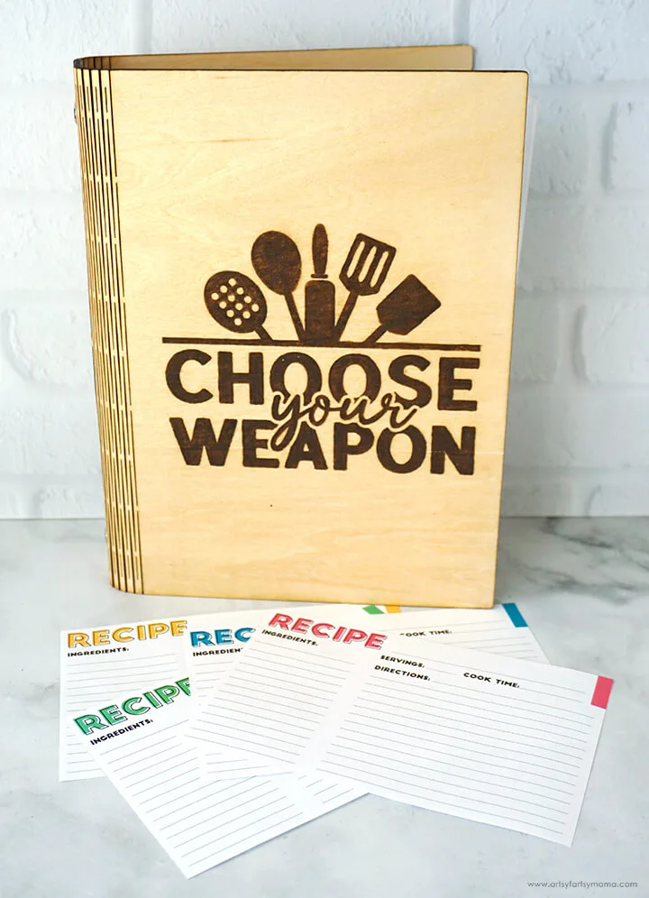 Wooden recipe binder with recipe cards in front that states choose your weapon with kitchen utensils engraved