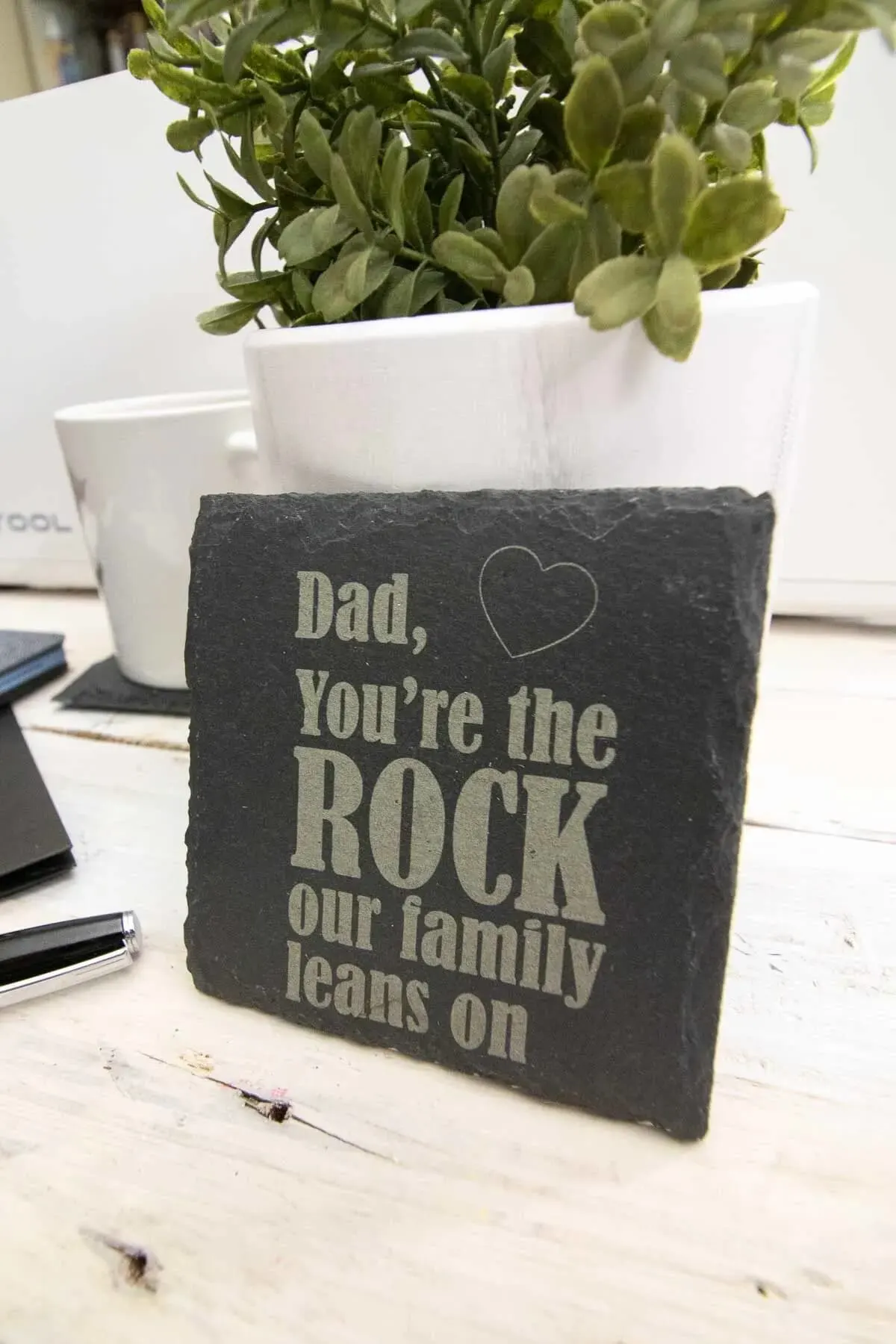 Slate coasters engraved with dad you're the rock our family leans on resting against a pot plant