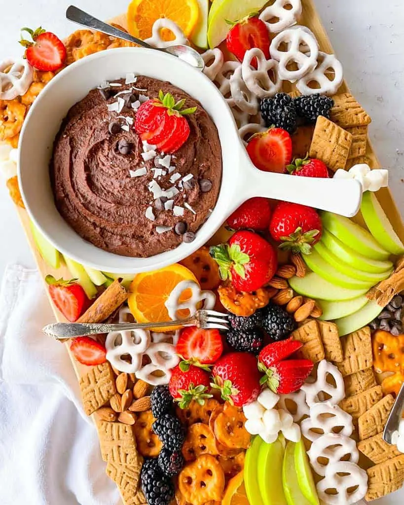 Large wooden serving board with healthy fruit snacks and chocolate treats with a chocolate hummus dip