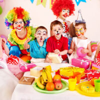5 kids at a birthday party with two clowns and a table of food including a tray of fresh fruit