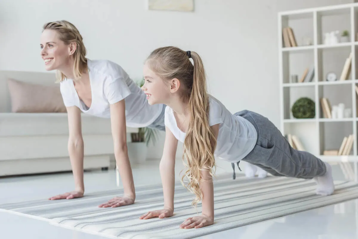 Mother and tween daughter doing yoga stretches in a large living room, wearing white tshirts and trackpants