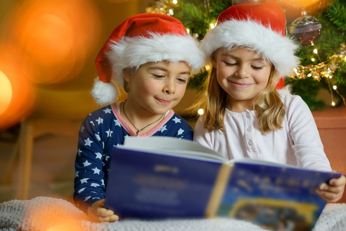 Two young girls with christmas hats reading a christmas book near the tree