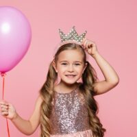 pretty little girl in fancy dress holding a pink balloon and putting on a crown headband.