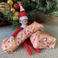 elf on the shelf with a party hat on holding birthday gifts under the christmas tree.