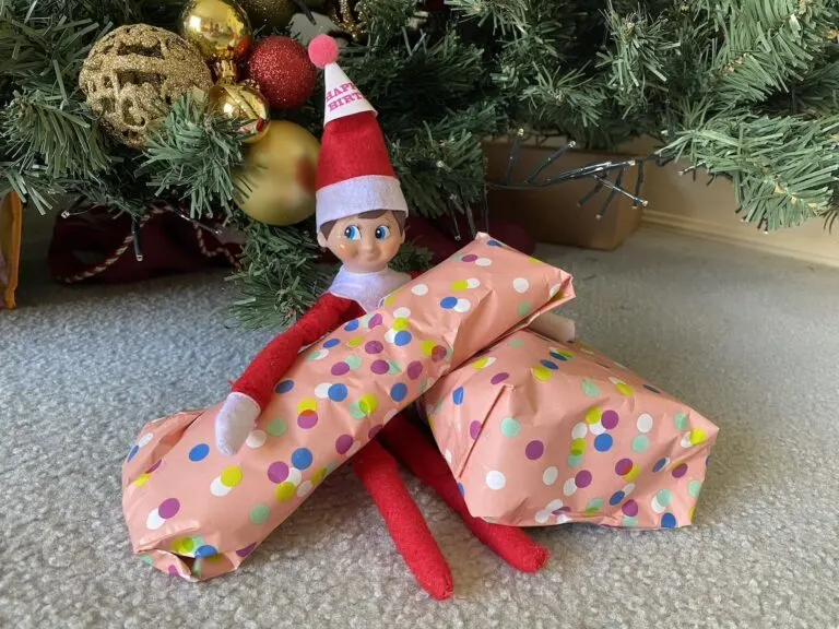 Elf on the shelf with a party hat on holding birthday gifts under the christmas tree.