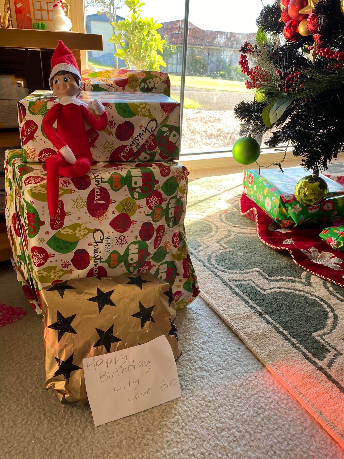 Elf on the shelf sitting on a stack of gifts with a birthday note.