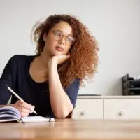 curly haired woman sitting at a table indoors reflecting and writing in a journal.