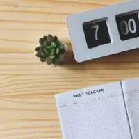 Top view or flat lay of habit tracker book, flip clock 7 am and succulent plant pot on wooden table background with copy space.